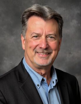 Bryce Gibbs, a middle-aged man with gray hair and a goatee, wearing a black blazer over a light blue shirt, poses confidently in front of a neutral gray studio background.