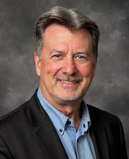 Bryce Gibbs, a middle-aged man with gray hair and a goatee, wearing a black blazer over a light blue shirt, poses confidently in front of a neutral gray studio background.