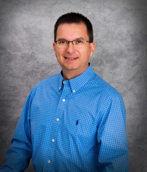 Jim Rein, wearing glasses and a blue checkered button-down shirt, stands in front of a gray textured background, smiling at the camera.