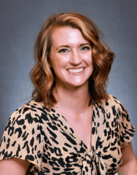 Woman with wavy brown hair smiles, wearing a leopard print top, reminiscent of Steve Pitman's bold style, against a gray background.