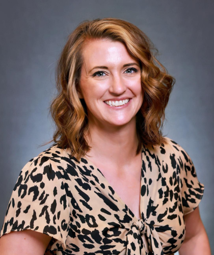 Woman with wavy brown hair smiles, wearing a leopard print top, reminiscent of Steve Pitman's bold style, against a gray background.