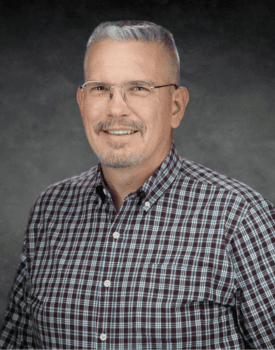 Steve Pitman, a man with short gray hair, glasses, and a goatee, smiles warmly while wearing a checkered shirt against a gray background.