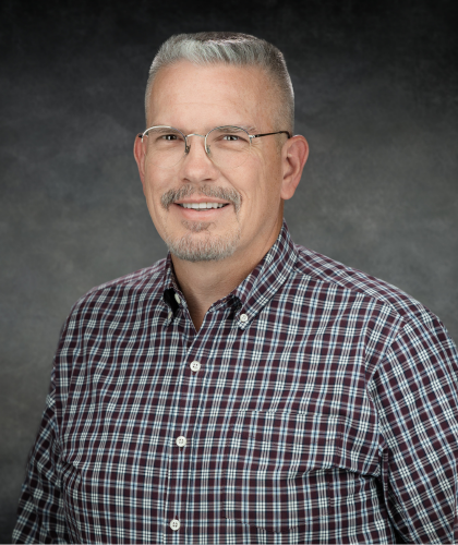 Steve Pitman, a man with short gray hair, glasses, and a goatee, smiles warmly while wearing a checkered shirt against a gray background.