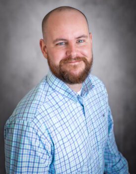 Brent Bright, a man with a beard and shaved head, wearing a light blue plaid shirt, smiles while seated in front of a gray background.
