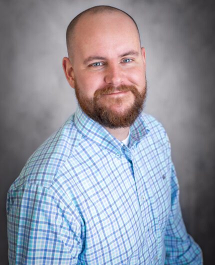 Brent Bright, a man with a beard and shaved head, wearing a light blue plaid shirt, smiles while seated in front of a gray background.