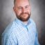 Brent Bright, a man with a beard and shaved head, wearing a light blue plaid shirt, smiles while seated in front of a gray background.