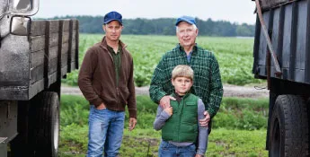 Three people stand between two trucks in a field: a young boy in a vest, an older man in a plaid shirt, and a younger man in a cap and brown jacket, all seemingly part of an animal ag operation.