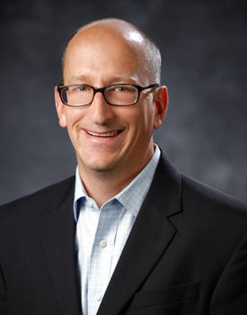 Brian Kuehl, A man wearing glasses smiles confidently, dressed in a black suit and light blue shirt, embodying the essence of Government and Public Affairs against a dark background.