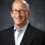 Brian Kuehl, A man wearing glasses smiles confidently, dressed in a black suit and light blue shirt, embodying the essence of Government and Public Affairs against a dark background.