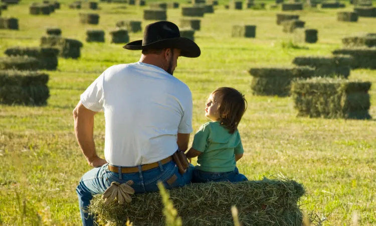 A man in a cowboy hat and a child sit on hay bales, surrounded by fields of commodity crops and scattered hay bales.