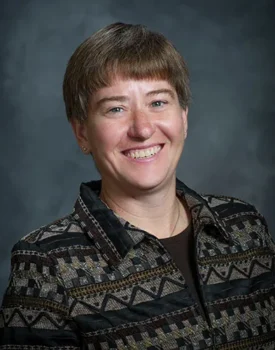 Donna Funk with short hair is smiling brightly, styled in a patterned jacket against a gray background, embodying the spirited essence of Donna Funk.