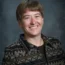 Donna Funk with short hair is smiling brightly, styled in a patterned jacket against a gray background, embodying the spirited essence of Donna Funk.