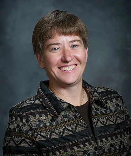 Donna Funk with short hair is smiling brightly, styled in a patterned jacket against a gray background, embodying the spirited essence of Donna Funk.