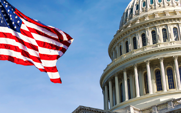 The U.S. flag waves proudly in front of the domed Capitol building in Washington D.C., a symbol of government and public affairs, under a clear blue sky.
