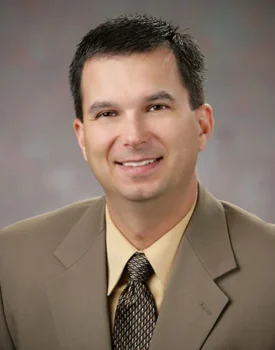 Jim Rein, A person wearing a brown suit, beige shirt, and patterned tie, exuding a next-gen confidence with a slight smile and short dark hair against a neutral backdrop.