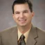 Jim Rein, A person wearing a brown suit, beige shirt, and patterned tie, exuding a next-gen confidence with a slight smile and short dark hair against a neutral backdrop.