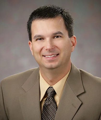 Jim Rein, A person wearing a brown suit, beige shirt, and patterned tie, exuding a next-gen confidence with a slight smile and short dark hair against a neutral backdrop.