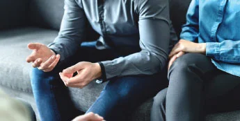 Two people are sitting on a couch, immersed in conversation. One's hands are gesturing animatedly, while the other rests a reassuring hand on their arm. Both are dressed in casual clothing, embodying the relaxed vibe of this next-gen connection.