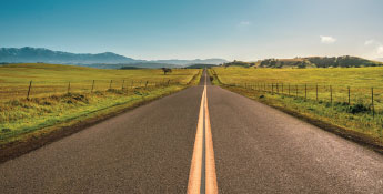 A straight, empty road stretches into the distance through a vast grassy landscape with distant mountains under a clear blue sky, offering a next-gen glimpse of untouched beauty and endless possibilities.