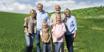 A next-gen family of six, including two children, stands together on a grassy field with a blue sky and trees in the background.