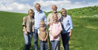 A group of six people, three adults and three children, stands together on a grassy field beneath a bright blue sky, embodying the spirit of the Next Gen.