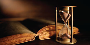 An hourglass with sand flows beside an open book on a dimly lit wooden surface, capturing a timeless moment that bridges the past with the Next Gen.