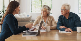 A woman is discussing Next Gen documents with an older couple at a wooden table. A laptop and coffee cups are also on the table in a bright room.