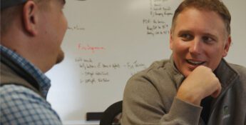 Two men sitting and talking in an office setting, with a whiteboard in the background covered in handwritten notes.