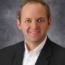 A smiling Thomas in a suit and white shirt stands confidently against a gray background, exuding the expertise of farm business management.
