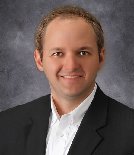A smiling Thomas in a suit and white shirt stands confidently against a gray background, exuding the expertise of farm business management.