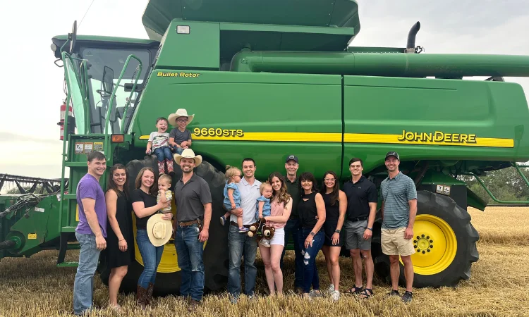 A group of people, including adults and children, stand in front of a large green John Deere farming vehicle in a field, embodying the Next Gen spirit. Some wear hats and casual clothing.