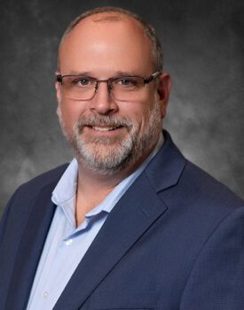 Headshot of Pinion Principal Tyson Shoemaker wearing glasses and a suit, with Gray background.