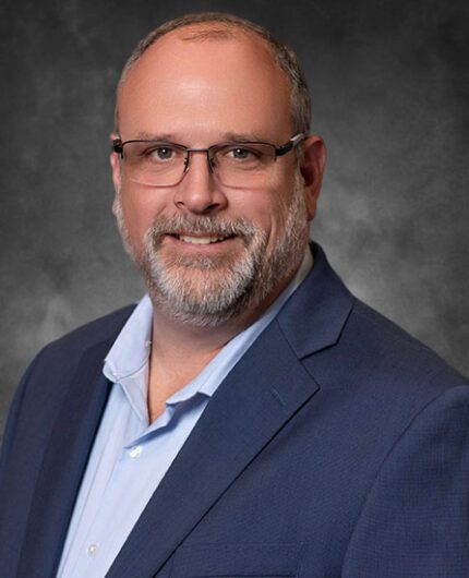 Headshot of Pinion Principal Tyson Shoemaker wearing glasses and a suit, with Gray background.