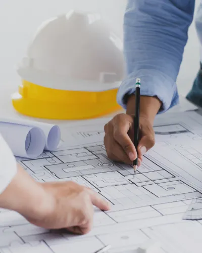 Two architects meticulously review architectural blueprints laid out on the table, with one hand pointing and another holding a pencil. In the background, a construction helmet and rolled-up plans hint at their ongoing project discussions.