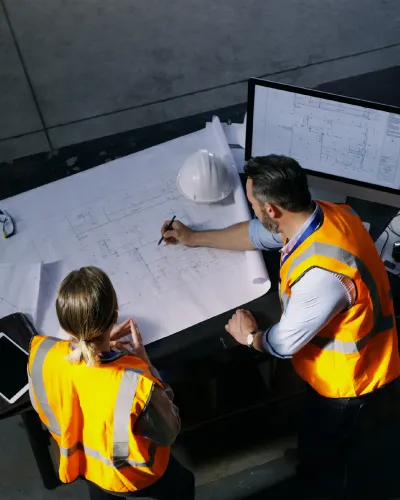Two construction workers in orange vests discuss blueprints on a table. Resembling architects at work, one points at the plans, while a hard hat and monitor displaying diagrams are nearby.