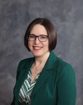 Baalman Lisa with a green blazer and patterned shirt, standing against a grey backdrop.