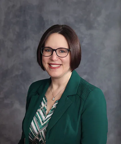 Baalman Lisa with a green blazer and patterned shirt, standing against a grey backdrop.
