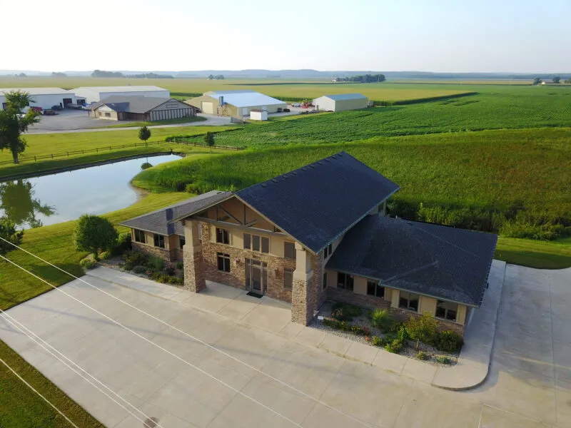 Aerial view of a large rural house with adjacent buildings, surrounded by farmland. A pond is visible to the left, and the landscape features open fields under a clear sky.
