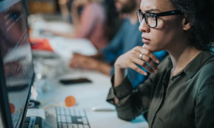 A person wearing glasses sits at a desk, deep in thought as they review IT consulting strategies on the computer monitor, hand resting on their chin, while colleagues work diligently in the background.