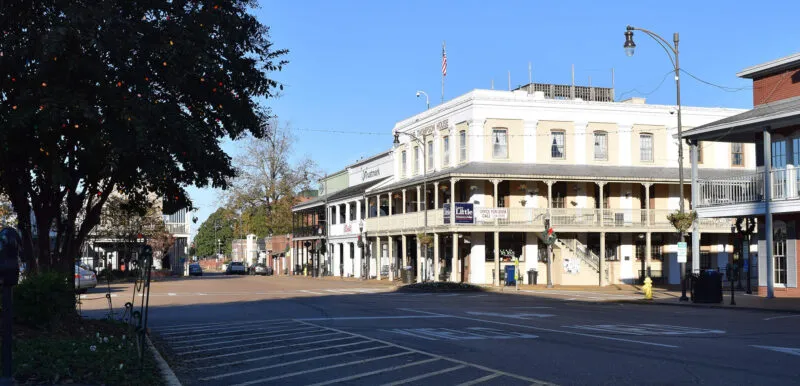 Street view of a historic district with an old-fashioned multi-story building, trees on the left, and a clear blue sky. The area features signage and streetlights.