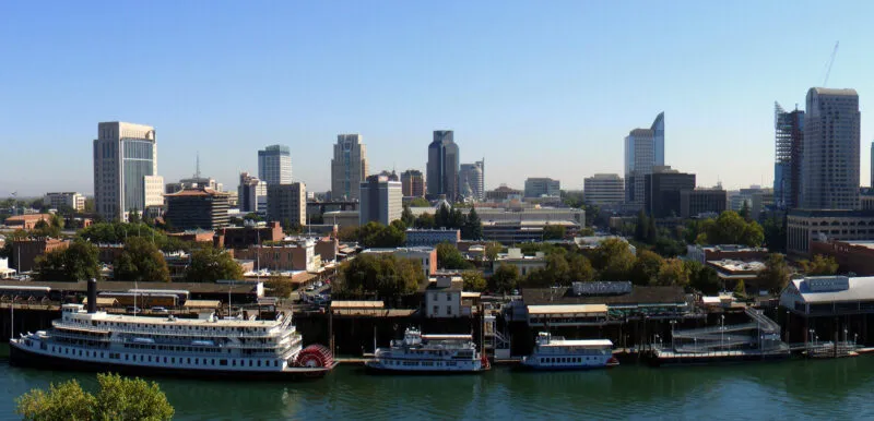 Panoramic view of a city skyline with modern buildings, trees, and a river in the foreground. Several boats are docked along the riverbank.