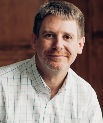 A man with short light brown hair and a trimmed beard, wearing a white checkered shirt, smiles slightly in front of a brown background, reflecting the approachable professionalism typical in IT consulting.