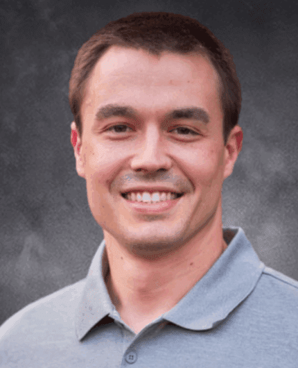Man with short brown hair wearing a light gray collared shirt, smiling in front of a dark, textured background—perfect for a professional profile in the commodity crops industry.