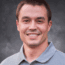 Man with short brown hair wearing a light gray collared shirt, smiling in front of a dark, textured background—perfect for a professional profile in the commodity crops industry.