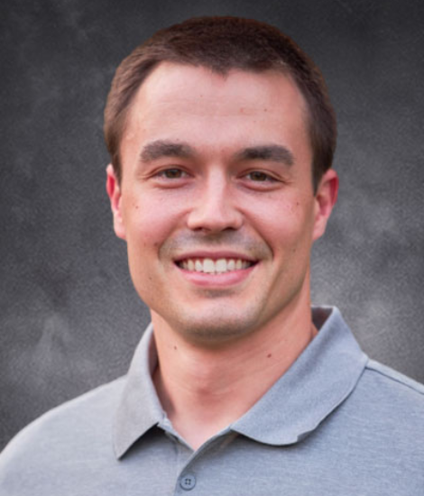 Man with short brown hair wearing a light gray collared shirt, smiling in front of a dark, textured background—perfect for a professional profile in the commodity crops industry.