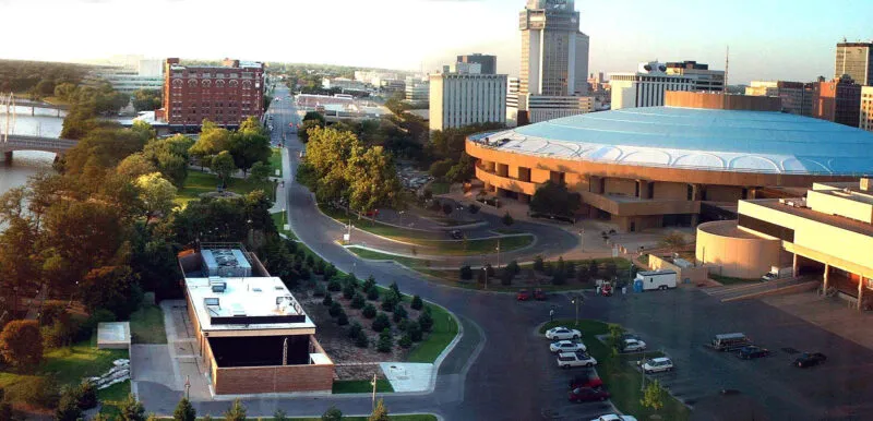 Aerial view of a cityscape featuring a large, round arena, several tall buildings, a river, and tree-lined roads.