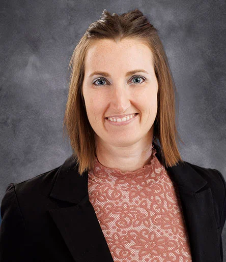 Melissa DeDonder, a woman with shoulder-length brown hair, smiles while wearing a black blazer over a lace-patterned pink top against a gray background.