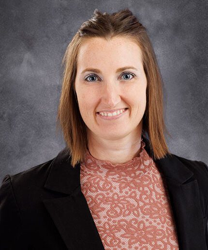 Melissa DeDonder, a woman with shoulder-length brown hair, smiles while wearing a black blazer over a lace-patterned pink top against a gray background.