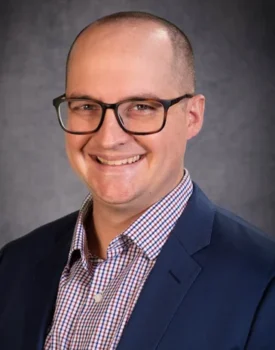 Alex Breitinger, wearing glasses, a checked shirt, and a dark blazer, smiles warmly against a neutral background.