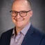 Alex Breitinger, wearing glasses, a checked shirt, and a dark blazer, smiles warmly against a neutral background.